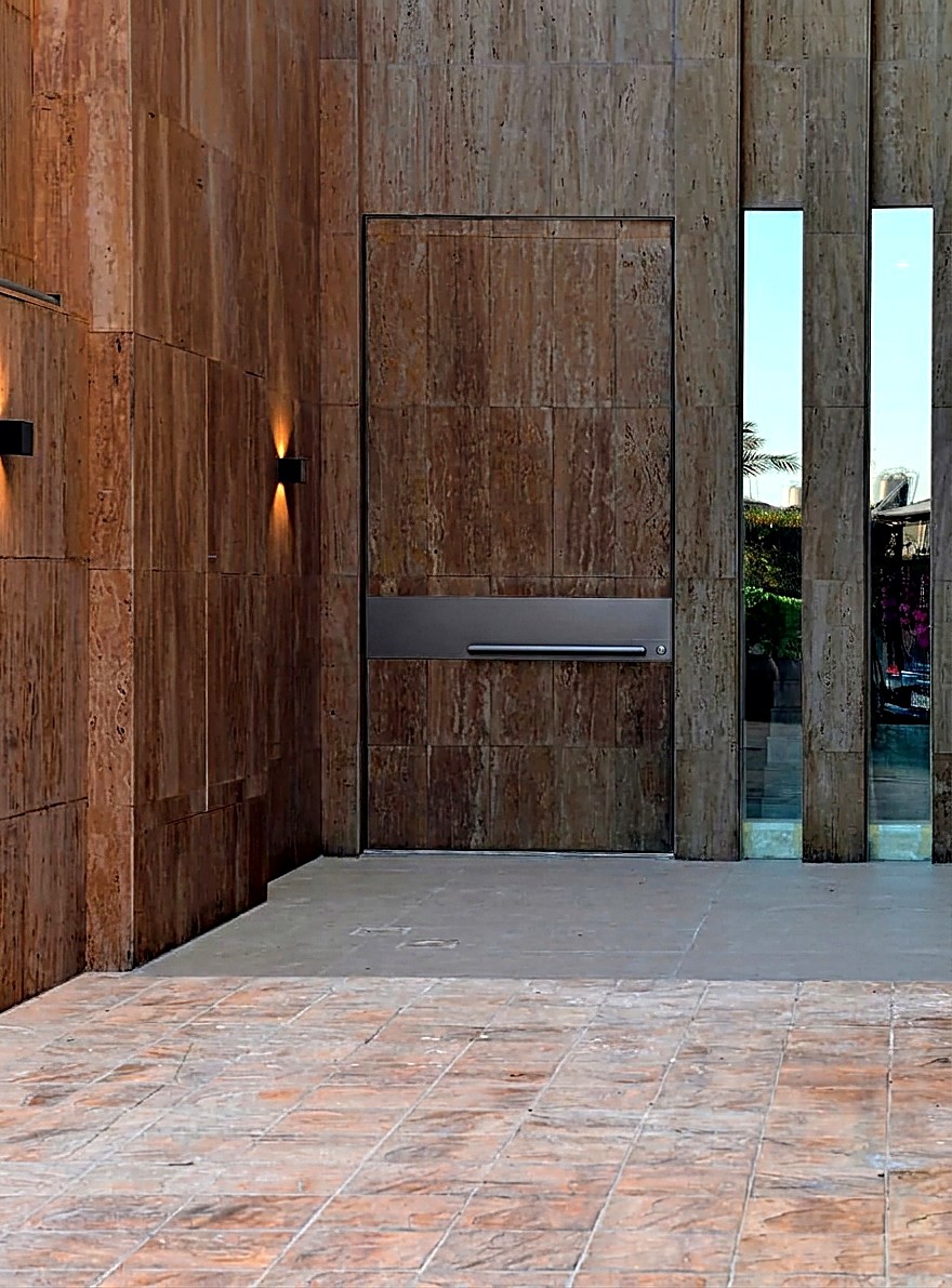 FORTIS entrance door clad in dark espresso travertine, fully flush with the stone facade, with a dark steel handle band