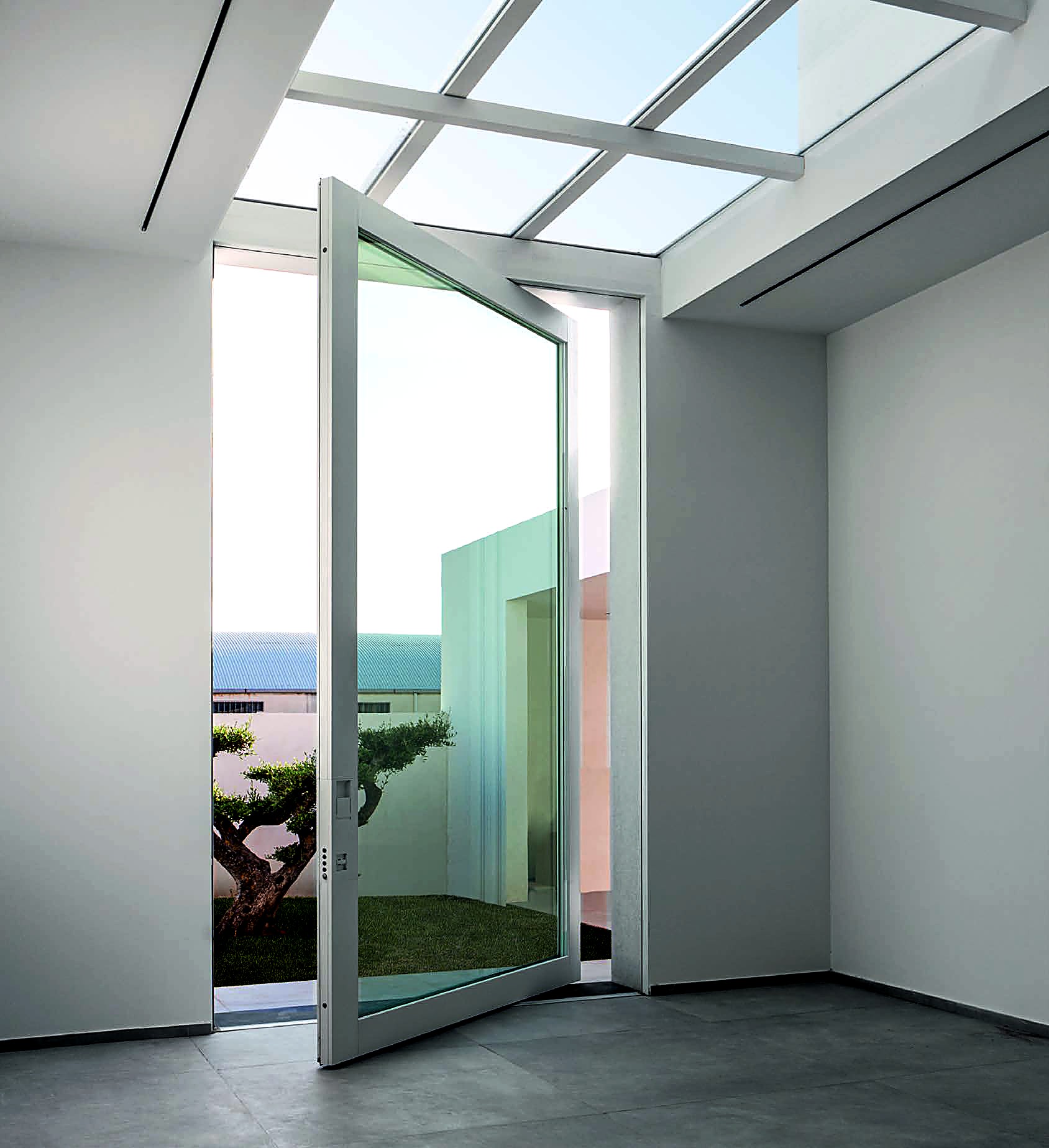 LUMEN glass pivot door in a slim white frame, open mid-pivot under a glazed skylight, olive tree in the courtyard beyond